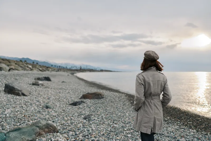 Woman in a gray coat and hat stands on a rocky beach looking at the sea under a cloudy sky. Concept for solitude, nature, and peaceful moments outdoors.
