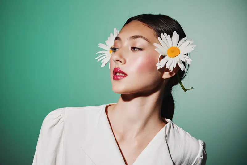 Young woman with wet hair and bright red lipstick wears white daisies as ear accessories on green background. Creative beauty concept combining fashion and floral style.