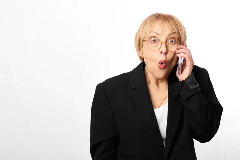 Elderly woman in business suit wearing glasses, shocked expression, holding smartphone to her ear, isolated on white background, communication concept