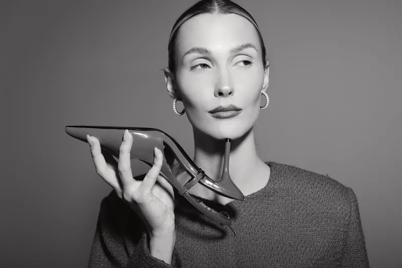 Portrait of a white slender woman holding a high heel shoe near her face, with slicked back hair in studio black and white.