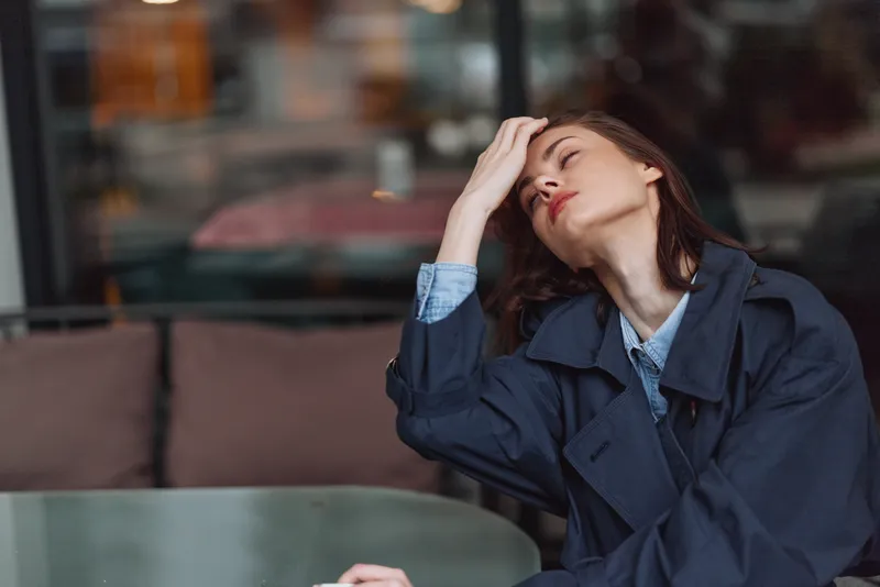 A young woman with brunette hair wearing a dark coat sits at a table indoors looking tired and stressed with her hand on her forehead and eyes closed.
