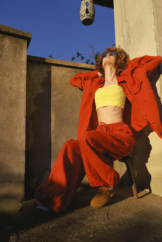 Confident woman in a red tracksuit and yellow crop top leans against a sunlit wall with arms raised, radiating bold, carefree energy and street style