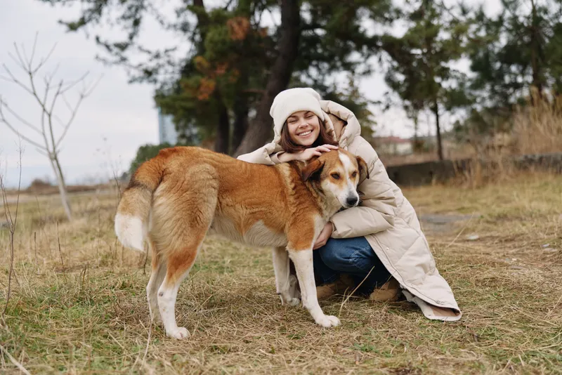 Woman in a beige coat hugs a dog in an open autumn field, trees in the background, a warm moment of companionship and outdoor activity with a beloved pet
