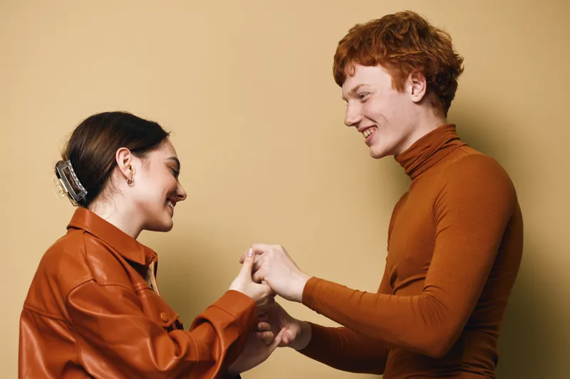 Couple, friendship and connection with smiling young man and woman holding hands for support and trust on beige background. Positive emotion and relationship between two people indoors.