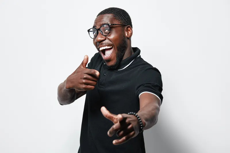 African American man with dark skin wearing glasses and black polo shirt smiling and pointing fingers at camera on white background. Happy young male with positive expression.