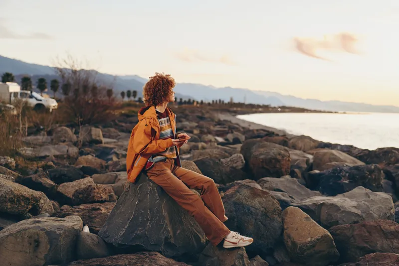 Woman lifestyle outdoor seated on rocks at seaside during sunset, casual jacket and pants, holding phone in hand and looking ahead, calm relaxation, warm evening light, travel mood