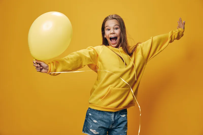 Girl smiling and holding yellow balloon with excitement in casual sweater and shorts, standing against yellow background for happiness and celebration.