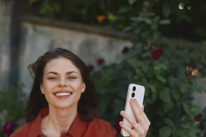 Vibrant lifestyle portrait of a smiling woman taking a selfie outdoors in a garden, wearing casual fashion, exuding friendly mood and confident expression while using a smartphone
