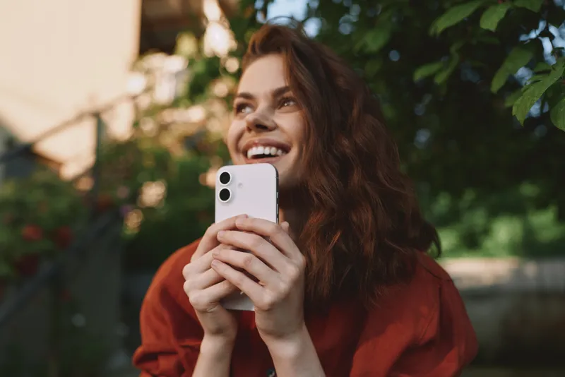 Bright woman lifestyle shot outdoors, smiling while holding a smartphone, casual red shirt, natural light, garden backdrop, candid moment of everyday joy