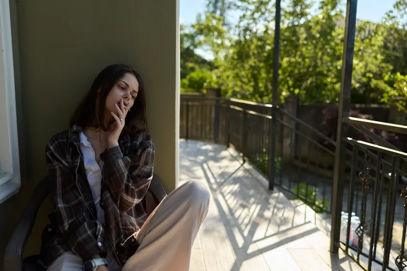 Woman sits on a balcony in casual clothing, smoking thoughtfully in soft sunlight, conveying a calm lifestyle moment on a sunny outdoor afternoon