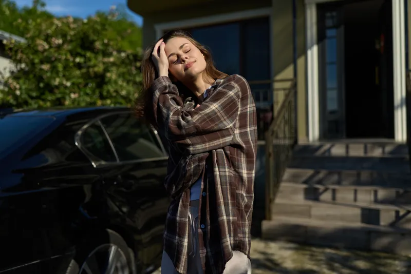 A woman in a plaid jacket poses outdoors beside a car, basking in sunlight with a relaxed smile. A casual lifestyle moment in a sunlit urban setting