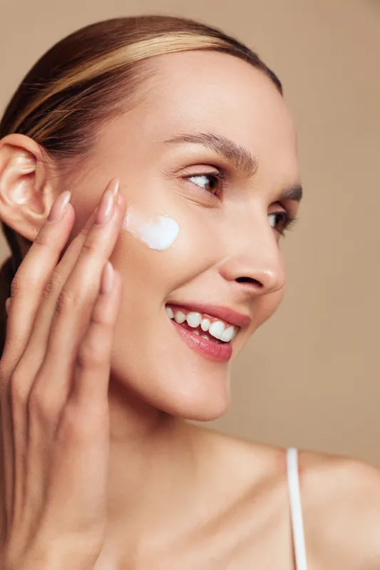 Cheerful woman applying cream to her cheek, closeup portrait, soft lighting, fresh skin, relaxed smile, skincare routine in a bright studio setting