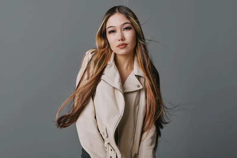 Young woman standing against gray background wearing beige jacket with long hairstyle falling over shoulders looking confidently at camera in studio portrait with soft lighting.