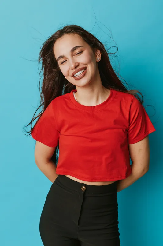 Young woman smiling with closed eyes wearing red crop top and black pants standing against blue background with hands behind back, long hair flowing and joyful expression.