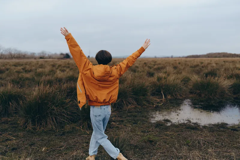 Woman outdoors lifestyle with raised arms in an open field near a small water path wearing a mustard jacket and jeans, her back to camera, calm nature moment under cloudy sky today
