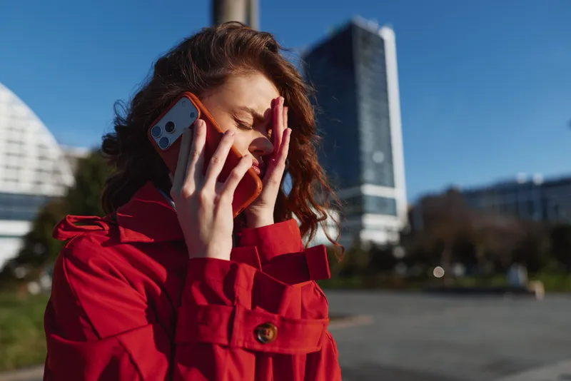 Young woman in a red coat talks on a smartphone outdoors while covering her face with her hand, expressing stress or frustration in an urban environment under clear sky.