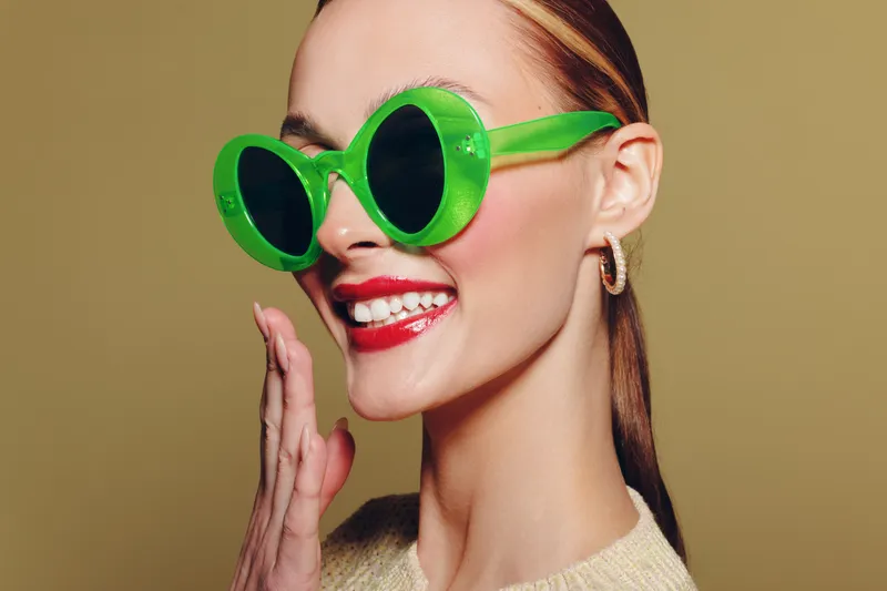 Smiling woman with green round sunglasses and red lipstick poses against beige background. Fashion portrait with trendy accessories and bright makeup. Concept of style and confidence.