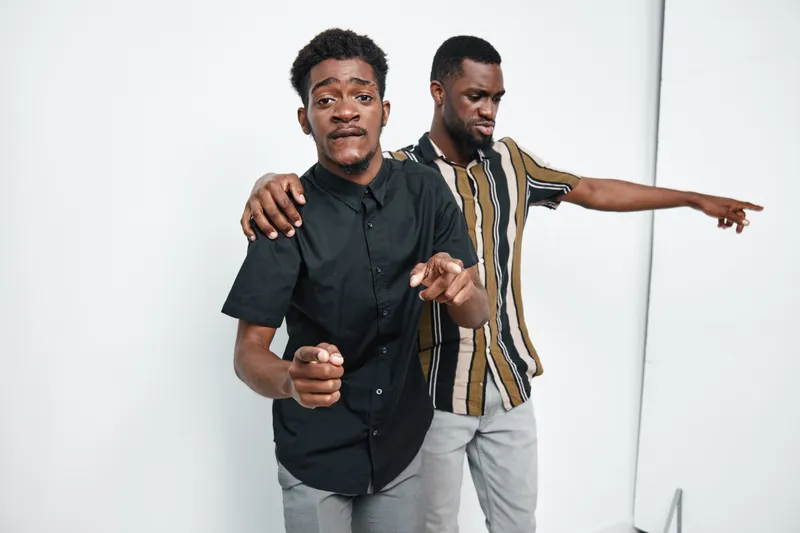 Two african men with dark skin posing together in casual clothes on white background. Friends, brothers or colleagues with one pointing and the other with arm on shoulder.