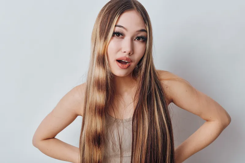 Young woman with long straight brown hair posing with hands on hips and surprised facial expression on neutral background. Beauty, hair care and female portrait studio shot.