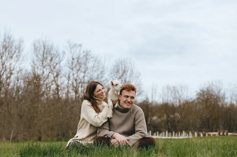 Young couple in cozy sweaters sits on grass in autumn park while woman holds a small white goat on her shoulder enjoying peaceful lifestyle moment together.