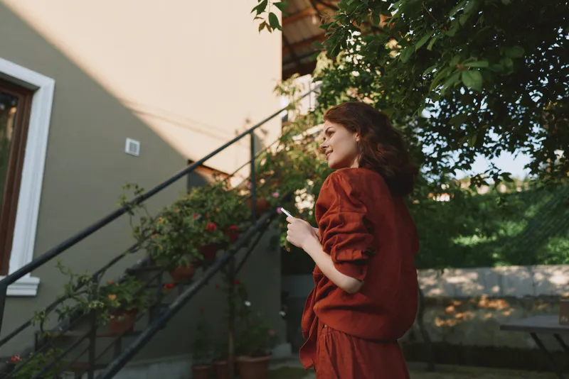 Smiling woman with wavy hair wearing a rust colored outfit stands beside an outdoor staircase. Casual lifestyle moment captured in warm daylight, conveying comfort, confidence, and everyday elegance