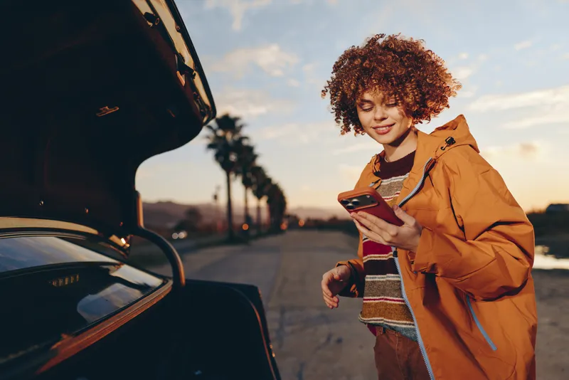 Woman lifestyle outdoors at sunset, smiling and walking beside car, holding smartphone, curly hair and warm jacket, relaxed posture, golden sky, casual travel scene, soft focus today