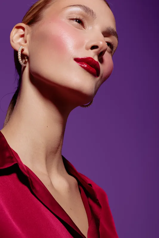 Close-up of a woman with glossy red lips and smooth skin wearing a red blouse against a purple background. Concept of beauty, makeup, fashion, and elegance.