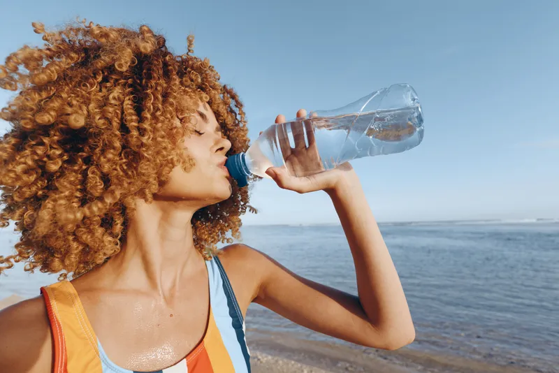 Woman curly hair drinking water from bottle by ocean outdoors, summer sunlight, hydration for wellbeing, breeze and blue sky, close up profile, refreshing healthy pause travel moment now