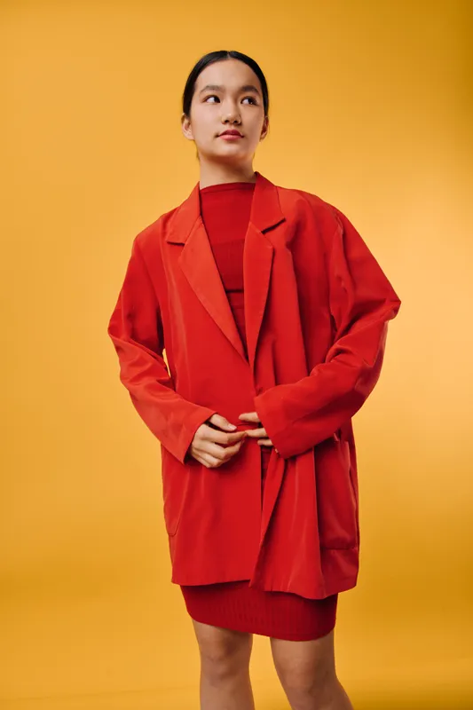 Woman in red oversized blazer and dress stands against yellow background, looking sideways with contemplative expression. Fashion, casual style and confident pose in studio portrait.