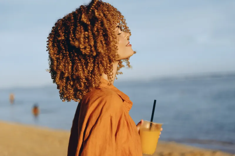 Young woman with curly hair enjoys a refreshing drink on a sunny beach while closing her eyes and feeling the gentle sea breeze in a relaxed moment.