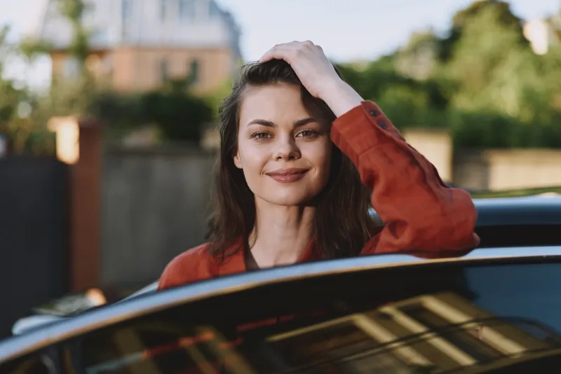A smiling woman leans on a car in a sunlit suburb, hand resting on her head. Natural lifestyle scene conveying everyday confidence and casual elegance