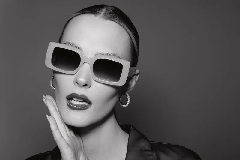 Stylish caucasian woman with pale skin wearing oversized sunglasses and hoop earrings poses with hand near face on grey background.