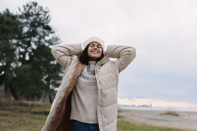 Woman outdoors wearing a warm coat and knit sweater, smiling with hands behind head, winter landscape and soft light, cozy fashion moment in chilly air and scenery.