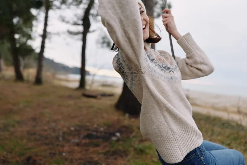 Woman on a swing outdoors at a beachside park, wearing a knitted sweater. Trees, sand and sea blurred in the background for a carefree moment of play and relaxation.