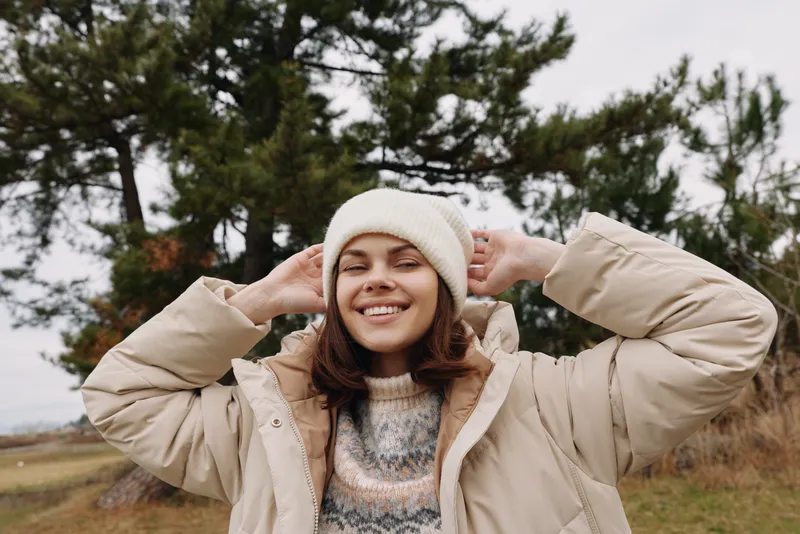 Woman in outdoor winter scene wearing a warm jacket and knit beanie, smiling and adjusting her coat, casual fashion portrait on a chilly day with natural scenery.