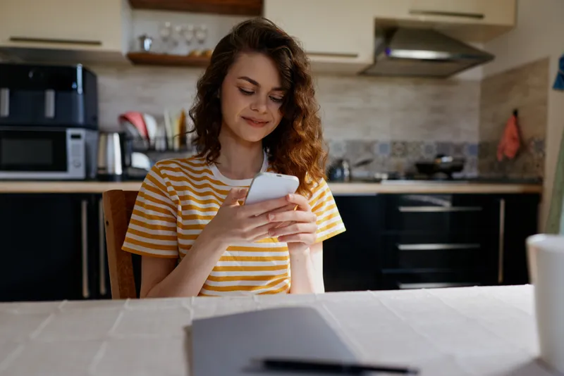 Smiling woman in a striped shirt using a smartphone at a kitchen table, candid home scene, casual lifestyle, everyday technology use, warm natural light, cozy kitchen atmosphere, relaxed mood