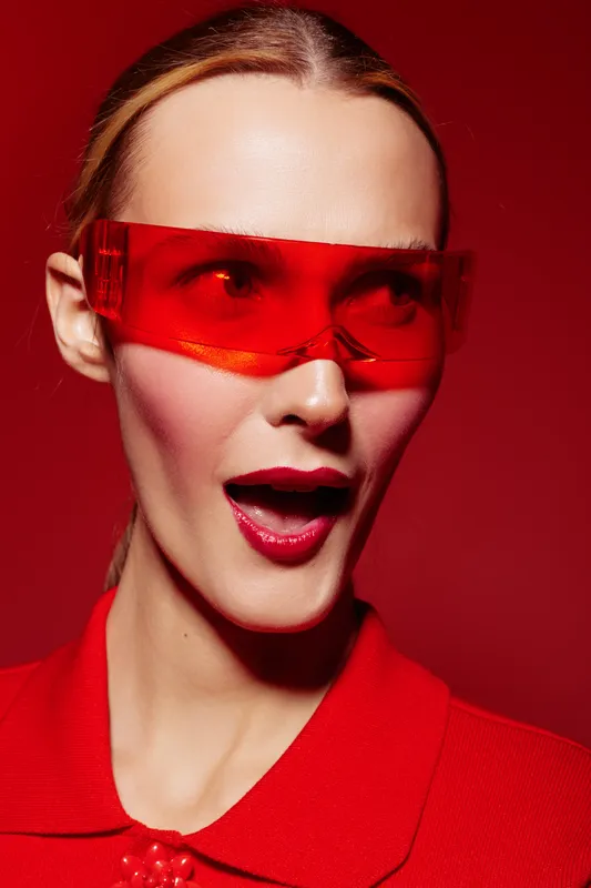 Young woman with red lips and red sunglasses looks surprised on red background. Fashion portrait with vibrant colors and modern style. Concept of beauty and bold makeup.