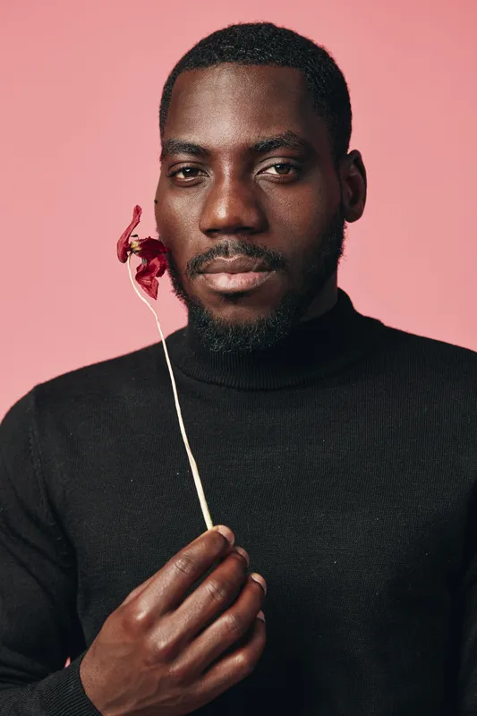 Black man with dark skin holding dried flower near face in creative conceptual visual style. Portrait with pink background and black turtleneck sweater showing calm expression and elegance.