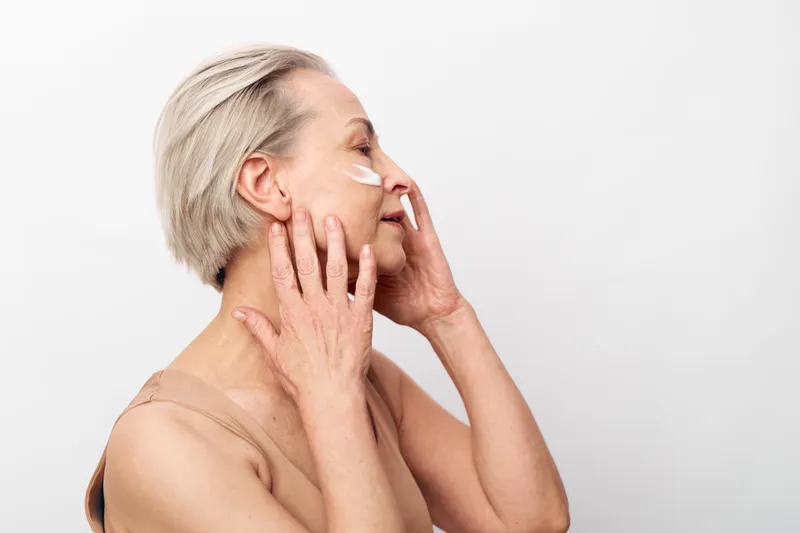 Mature woman with short grey hair applies white facial cream on cheeks, eyes closed, enjoying skincare routine. Concept for anti aging, self care, beauty, and natural skin health.