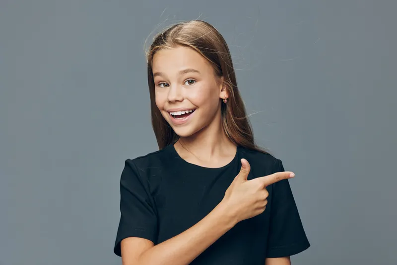 Girl teenager with long hair in black shirt smiling and pointing to side with hand isolated on gray background for youth and casual style.