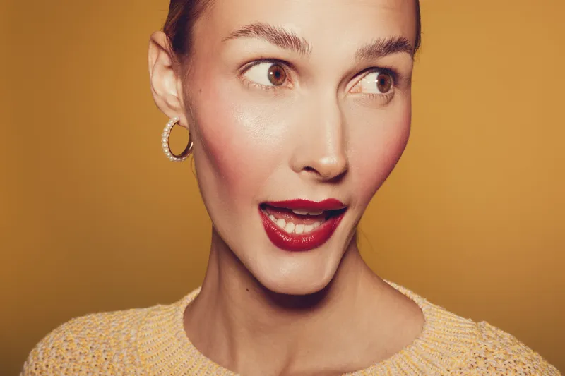Close-up of surprised woman with red lipstick and hoop earrings looking sideways on yellow background. Concept of emotion, beauty, fashion, and expression.