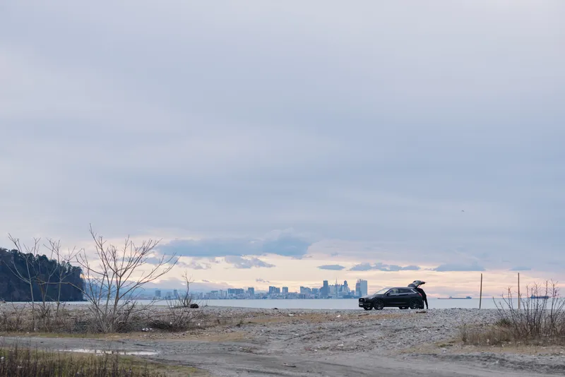 Car on a sandy shore at dusk with calm water and distant city skyline, empty beach, moody clouds, and sparse vegetation framing the horizon for a dramatic travel scene