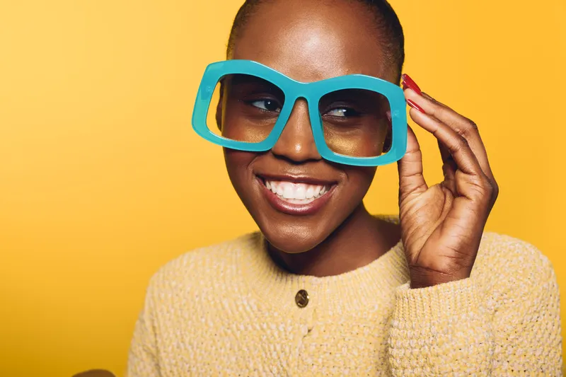 Happy African American woman with dark skin wearing large blue glasses smiles joyfully on yellow background. Concept of positive mood, beauty, fashion, and confident lifestyle portrait.