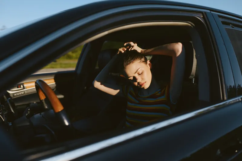 Young woman rests her head against the car window in a parked vehicle, wearing a striped top and warm sunlight streaming inside.