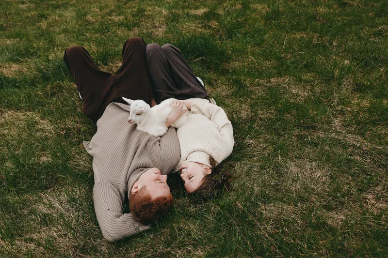 A couple rests on grass, smiling and holding a white goat kid between them. A candid lifestyle moment showing simple countryside joy, connection, and animal companionship.