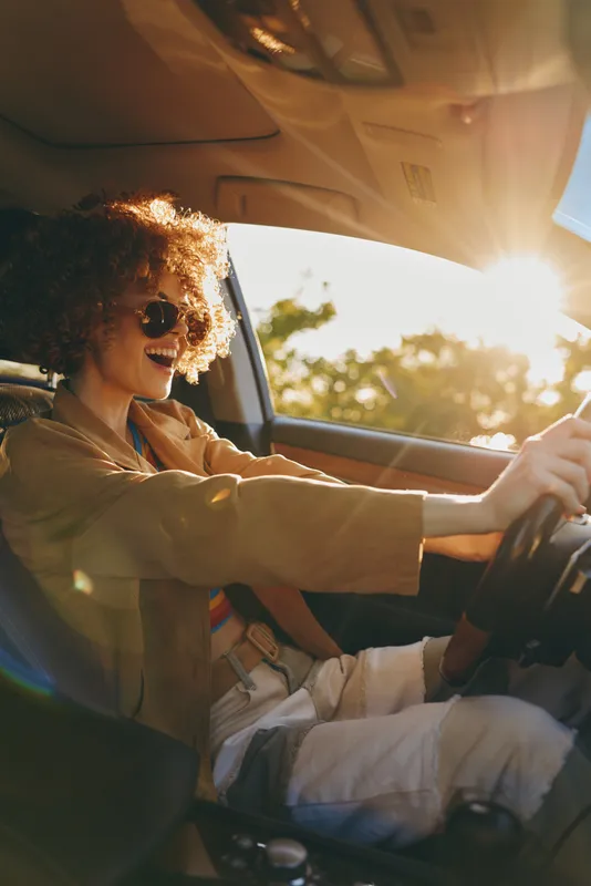 Smiling woman with curly hair driving a car, sunglasses on, warm retro tones, daylight through window, casual lifestyle and travel vibe, confident mood Bright sun glow adds nostalgia
