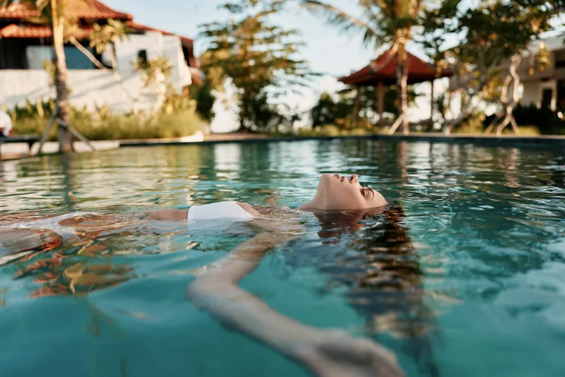 Relaxed woman floats in a calm pool at a tropical resort, enjoying warm sun, gentle water, and a peaceful moment among palm trees.