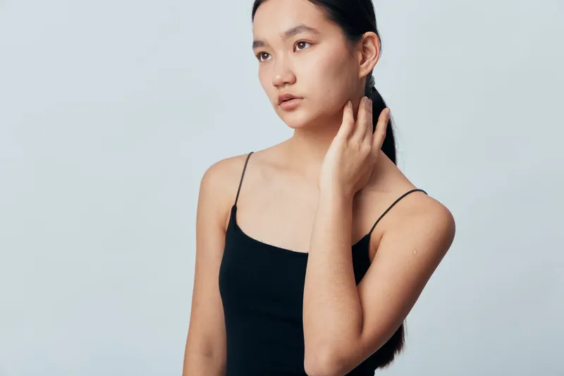 Young woman with long dark hair wearing black sleeveless top poses with hand near neck on light background. Natural beauty, contemplative expression and minimalistic style portrait indoors.