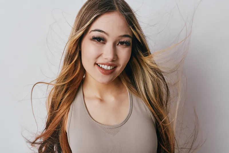 Young woman with long hair smiling at camera wearing casual top, natural makeup and happy expression with wind blowing strands in studio with white background.