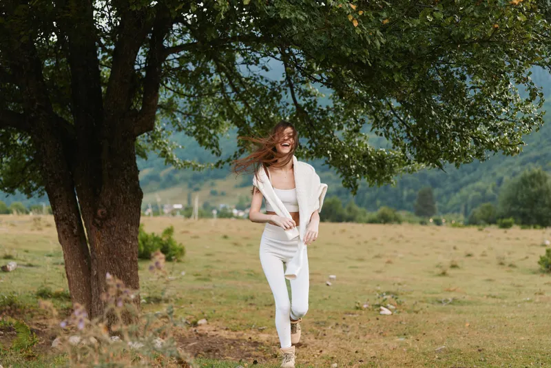 A young woman in white clothes runs happily near a large tree in a green field with mountains in the background. Lifestyle concept of freedom and nature.
